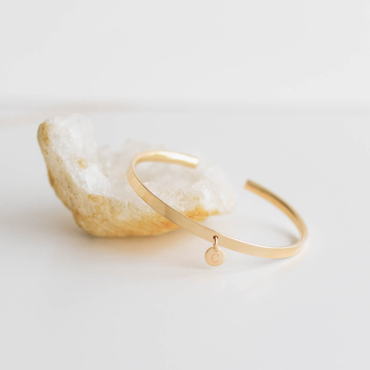 A minimalist gold-plated open cuff bracelet with a small circular charm engraved with the letter 'C', displayed on a clean white background.