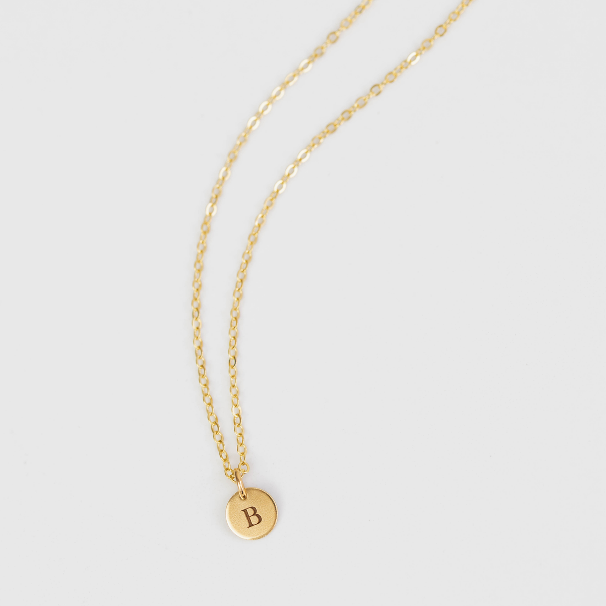 Gold necklace with a round pendant featuring the letter 'B' on a light gray background