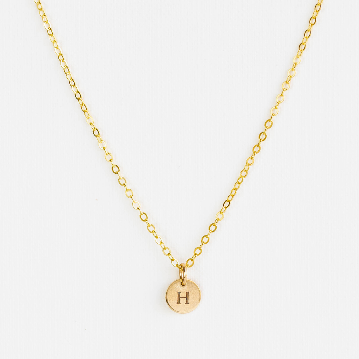 Gold necklace with a round pendant featuring the letter 'H' on a white background