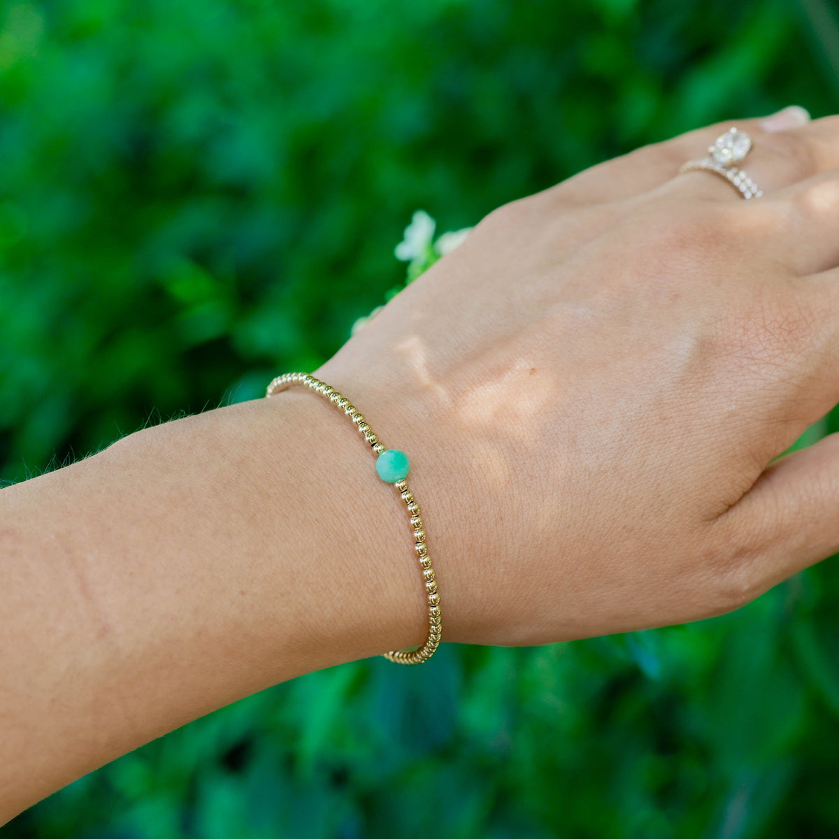 Hand wearing a gold bracelet with a green bead against a blurred green background