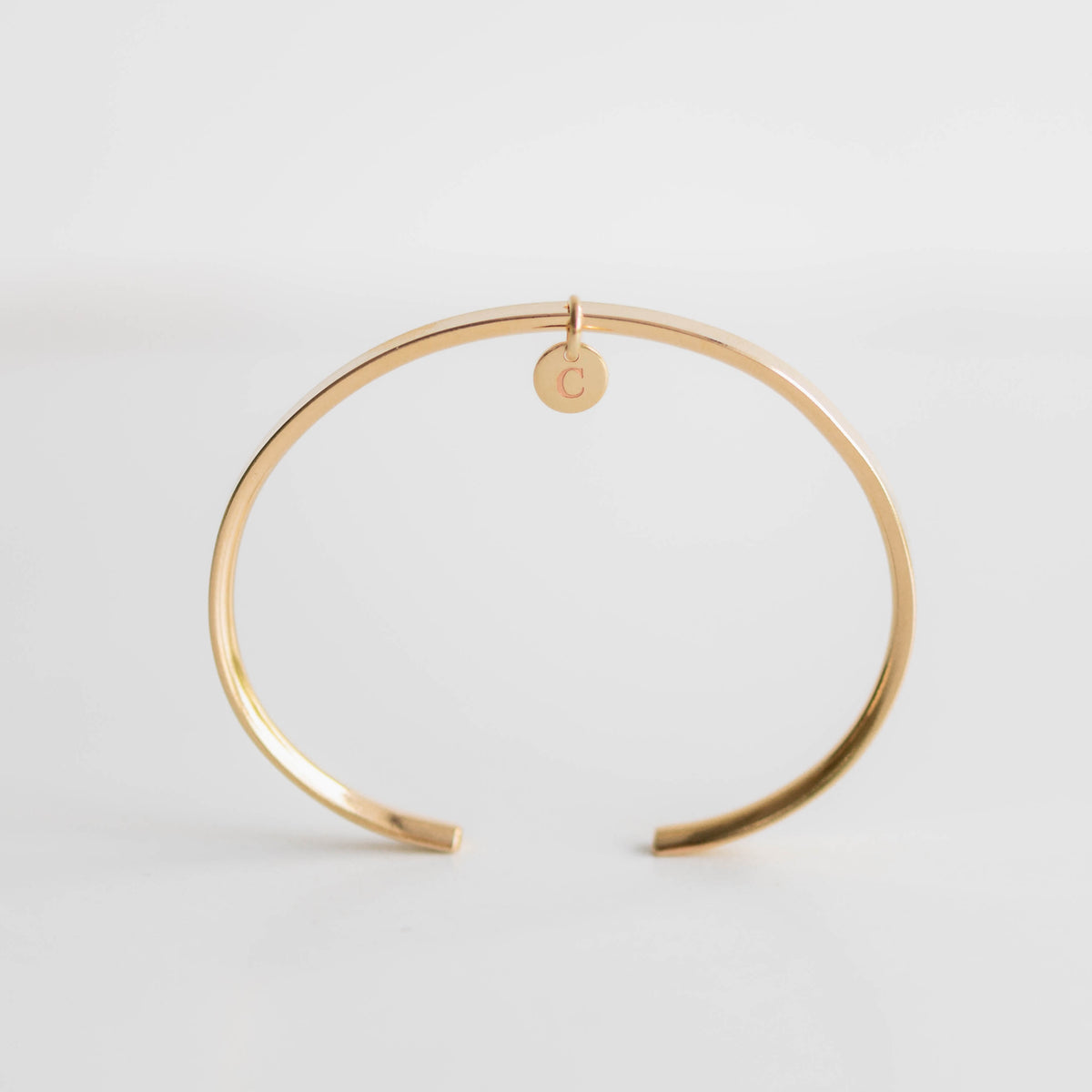 A minimalist gold-plated open cuff bracelet with a small circular charm engraved with the letter 'C', displayed on a clean white background.