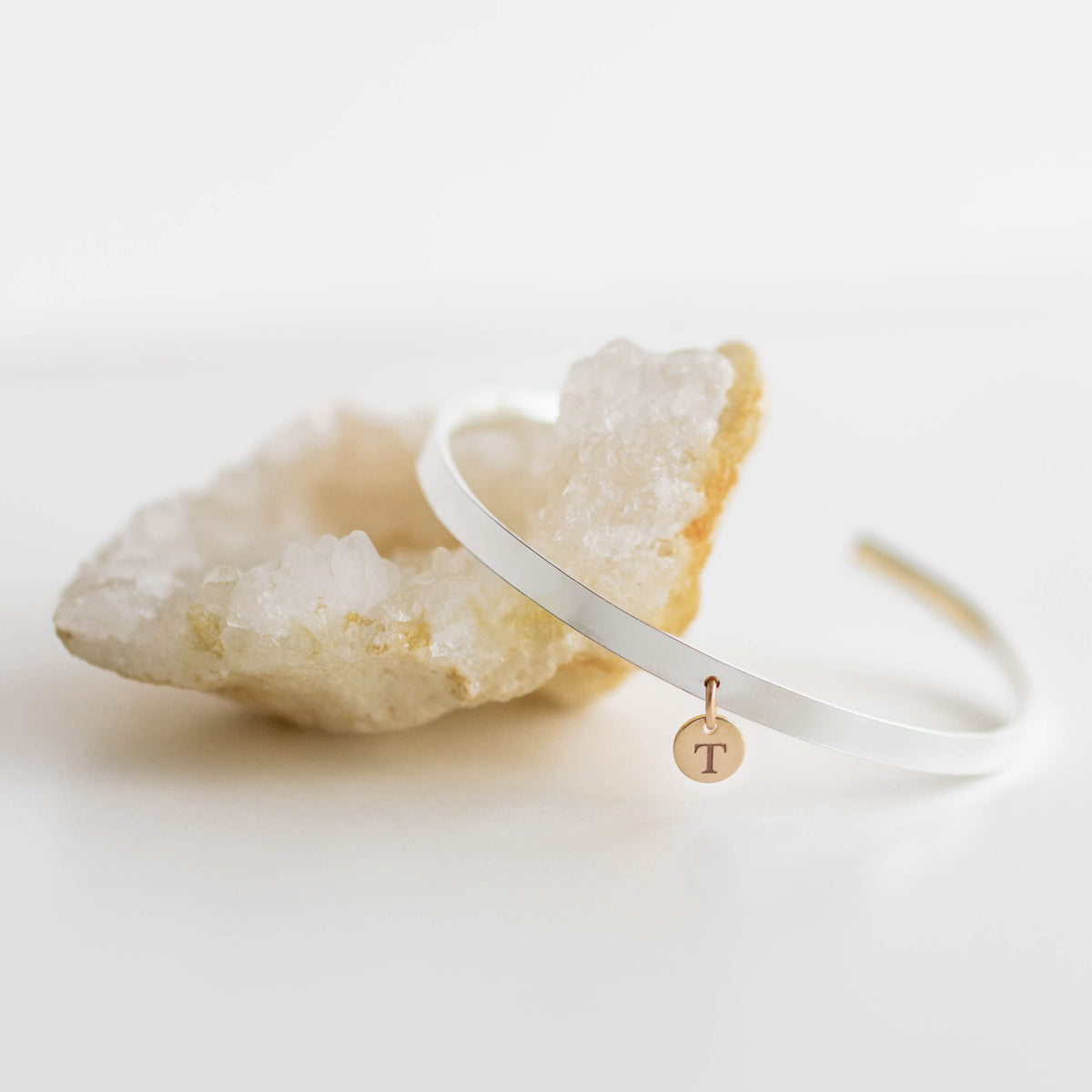A thin silver-tone open cuff bracelet with a small round rose-gold initial pendant engraved with the letter 'T', displayed on a white surface next to a natural crystal geode.