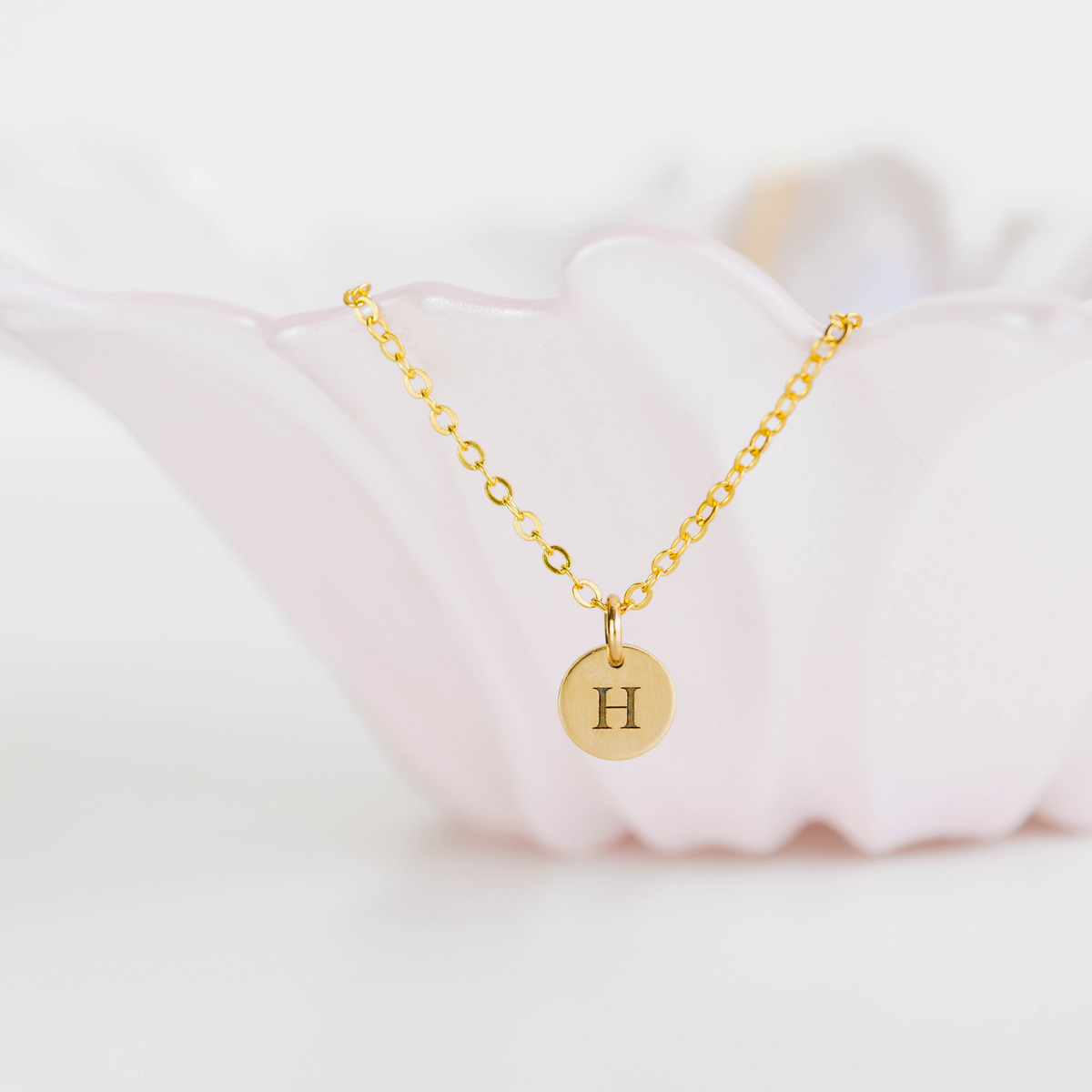 Gold necklace with a round pendant featuring the letter 'H' on a soft pink fabric background