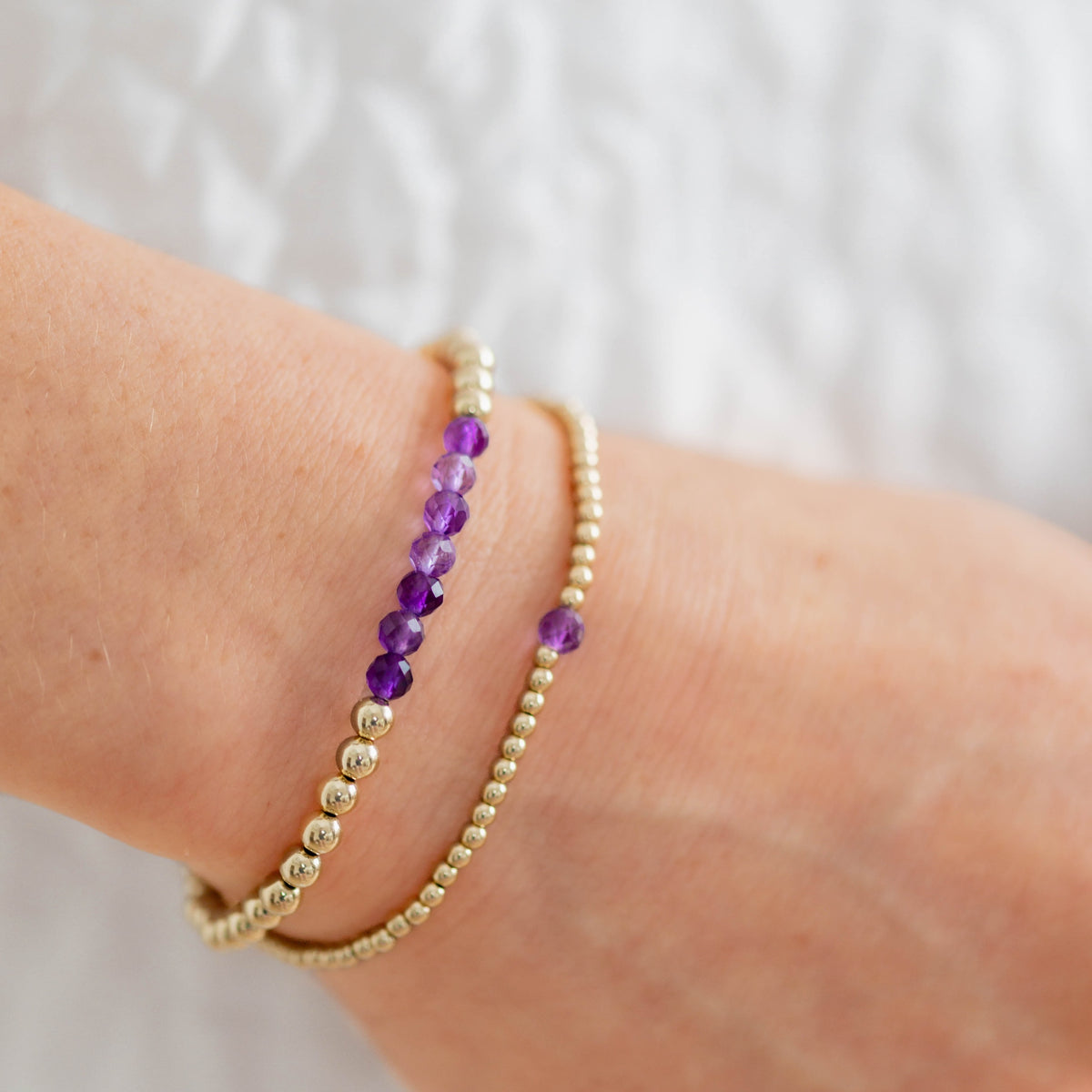 Two bracelets on a wrist, one gold and one with purple beads, against a neutral background.